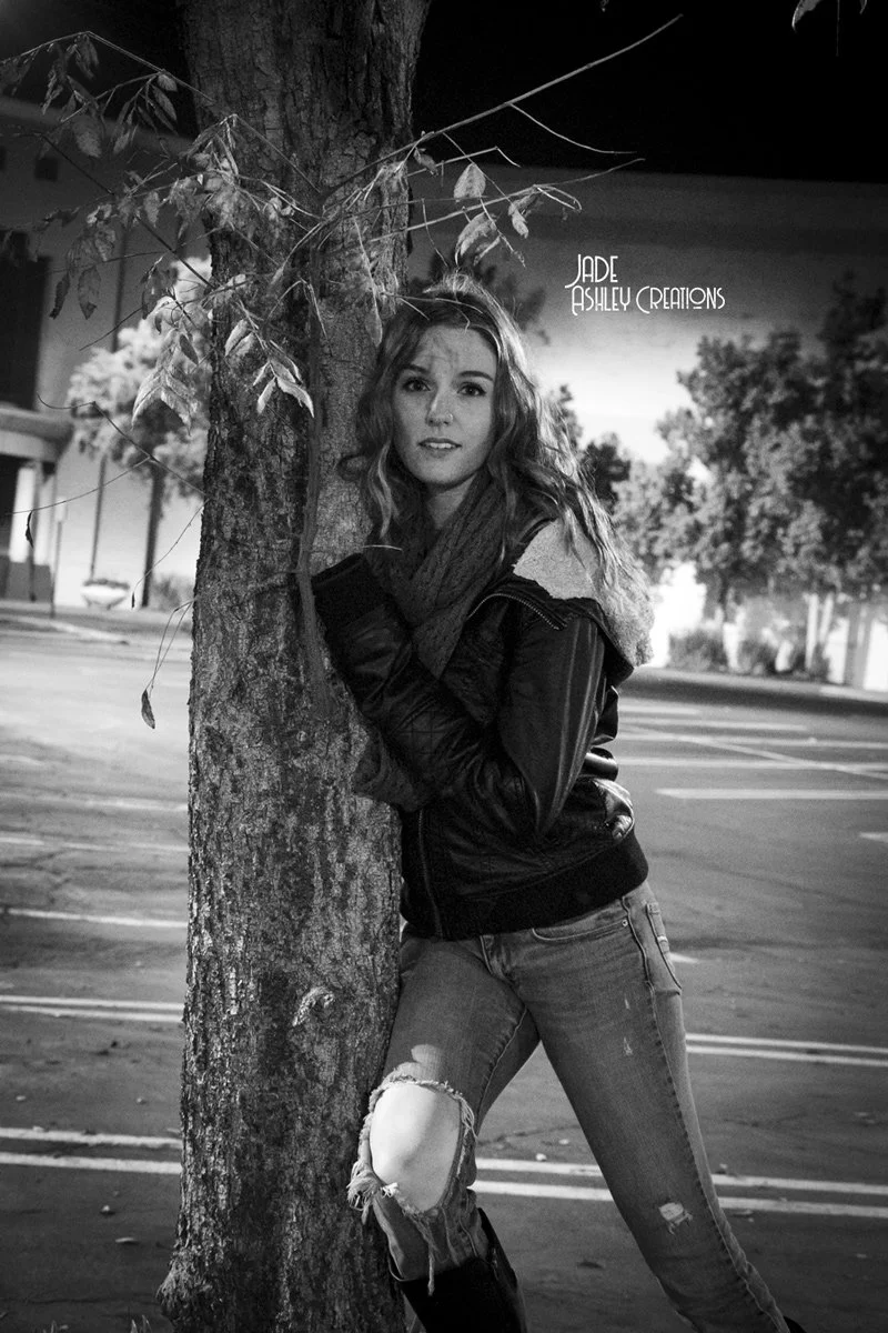 A young woman with wavy hair, wearing a leather jacket, scarf, and ripped jeans, stands outdoors at night leaning against a tree.