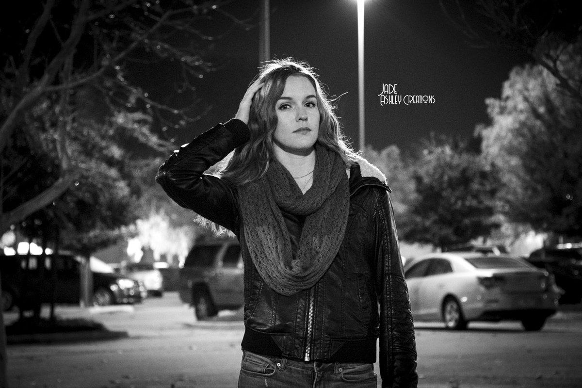 A young woman with wavy hair stands in a parking lot at night, wearing a leather jacket and a knitted scarf, with one hand touching her hair, illuminated by a streetlight.
