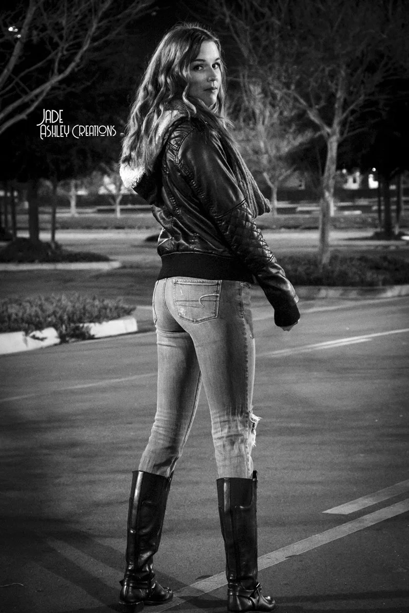 A young woman standing in a parking lot at night, wearing a leather jacket, jeans, and tall boots, looking over her shoulder at the camera.