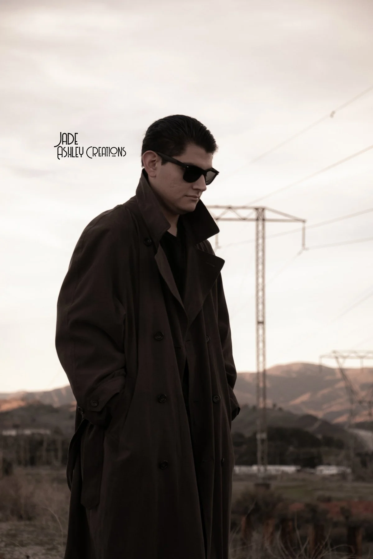 A man wearing sunglasses and a long dark trench coat standing outdoors with electrical towers and hills in the background, looking down.