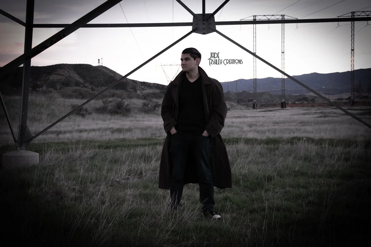 A man standing outdoors in a field under power lines and a large metallic structure, with mountains and utility poles in the background during dusk or dawn.