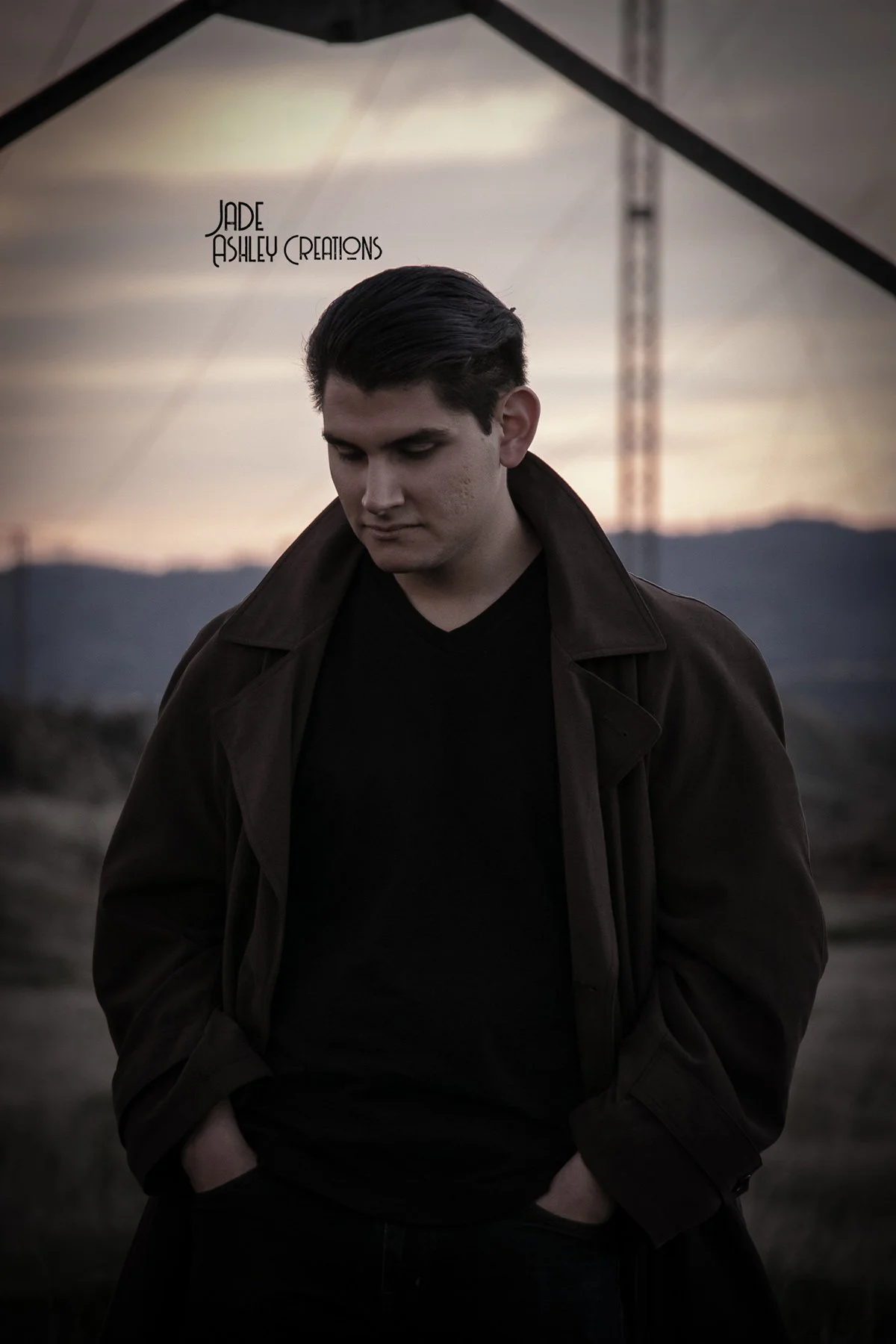 A young man in a dark brown coat and black shirt standing outdoors during sunset with mountains and a transmission tower in the background.