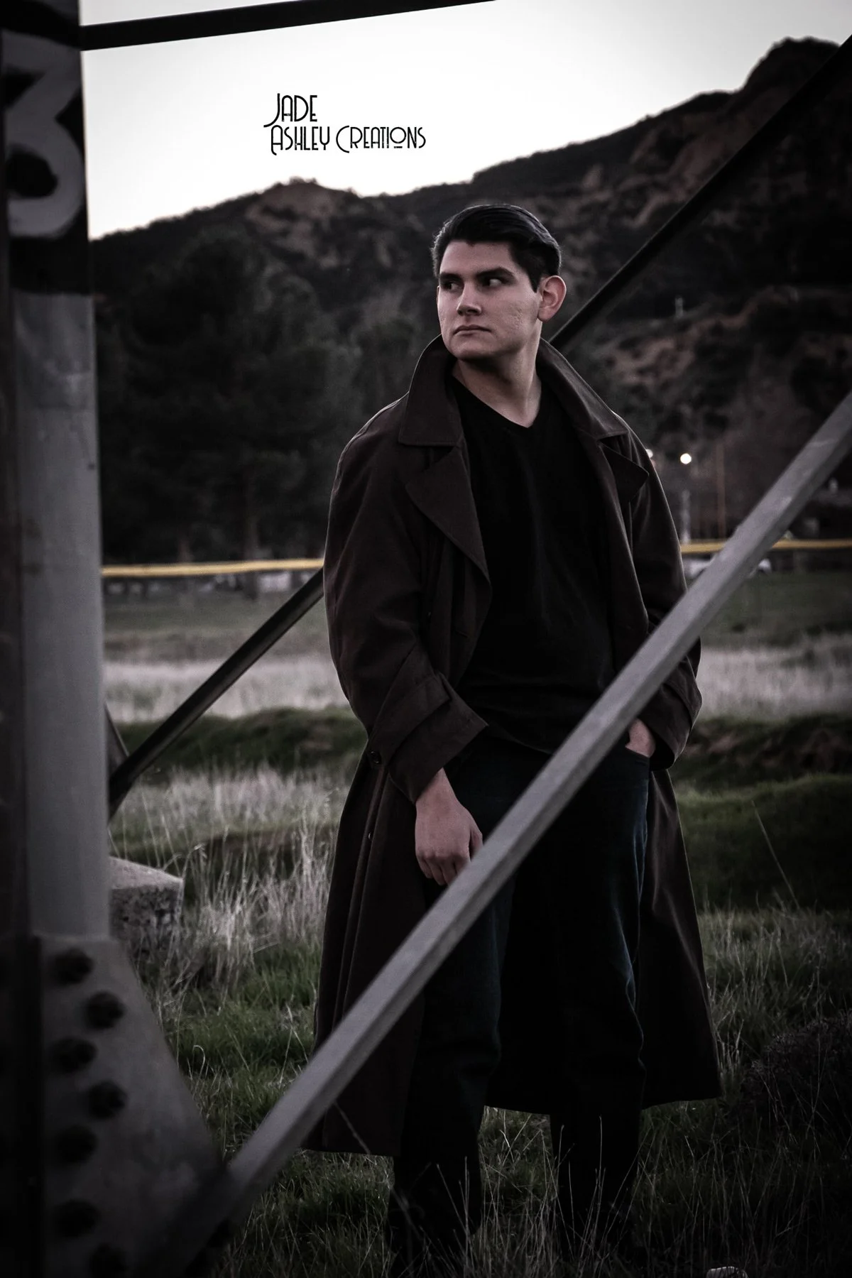 A young man with dark hair and light skin stands outdoors near a metal structure, wearing a long brown coat and black clothing, with a mountain landscape in the background during dusk.