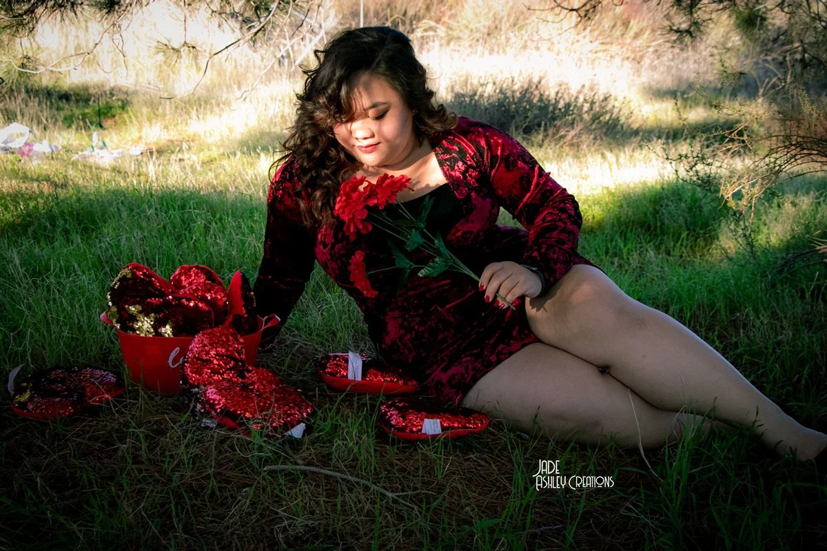 A woman with curly hair in a red velvet dress sitting on grass in a park, holding red flowers, surrounded by Valentine's Day decorations and gifts.
