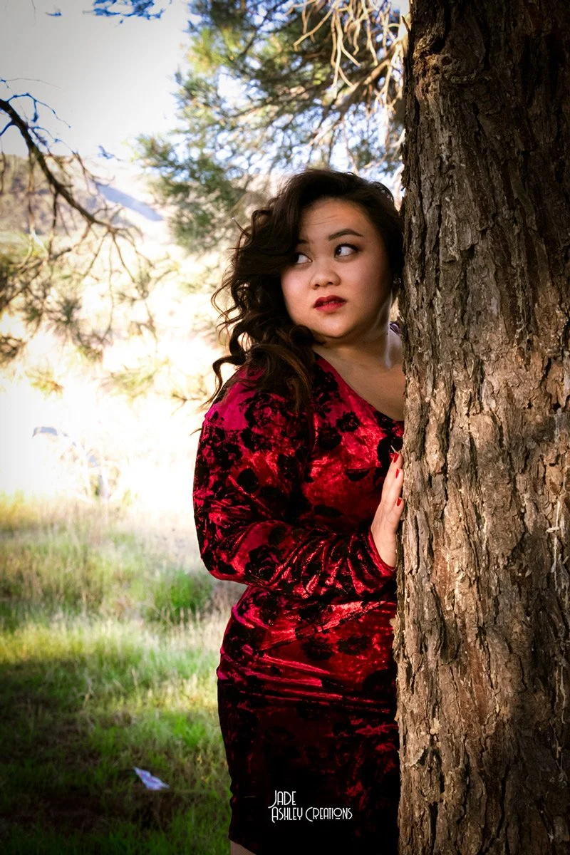 A woman with dark, curly hair peeking out from behind a large tree trunk in an outdoor park or forest setting. She is wearing a red velvet dress with black patterns and has red lipstick.