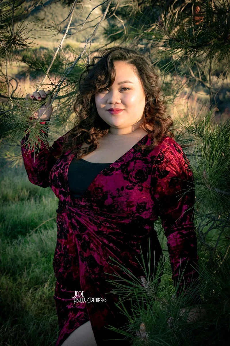 A woman with curly hair and light skin standing outdoors, surrounded by greenery and pine trees, wearing a red velvet dress with black accents, smiling at the camera.