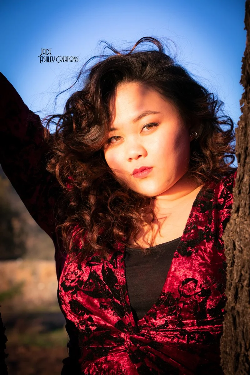 A woman with curly dark hair posing outdoors between two trees during sunset, wearing a black top and red velvet jacket with black floral patterns.