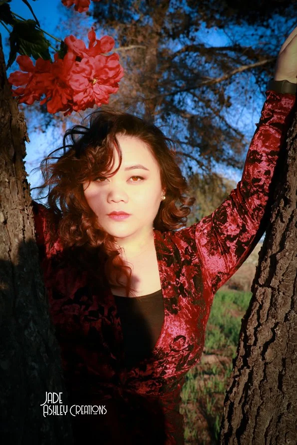 A woman with curly dark hair and light skin poses between two trees with red flowers, wearing a red velvet dress with black accents, during sunset with a blue sky in the background.