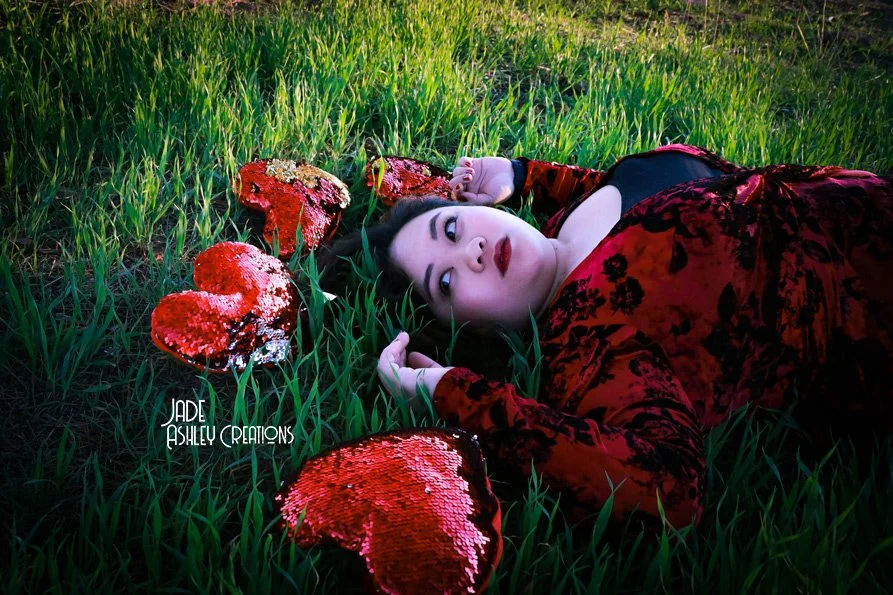 A woman with dark hair and makeup lying in tall green grass, surrounded by red sequined hearts. She wears a red velvet dress and looks directly at the camera with a calm expression.
