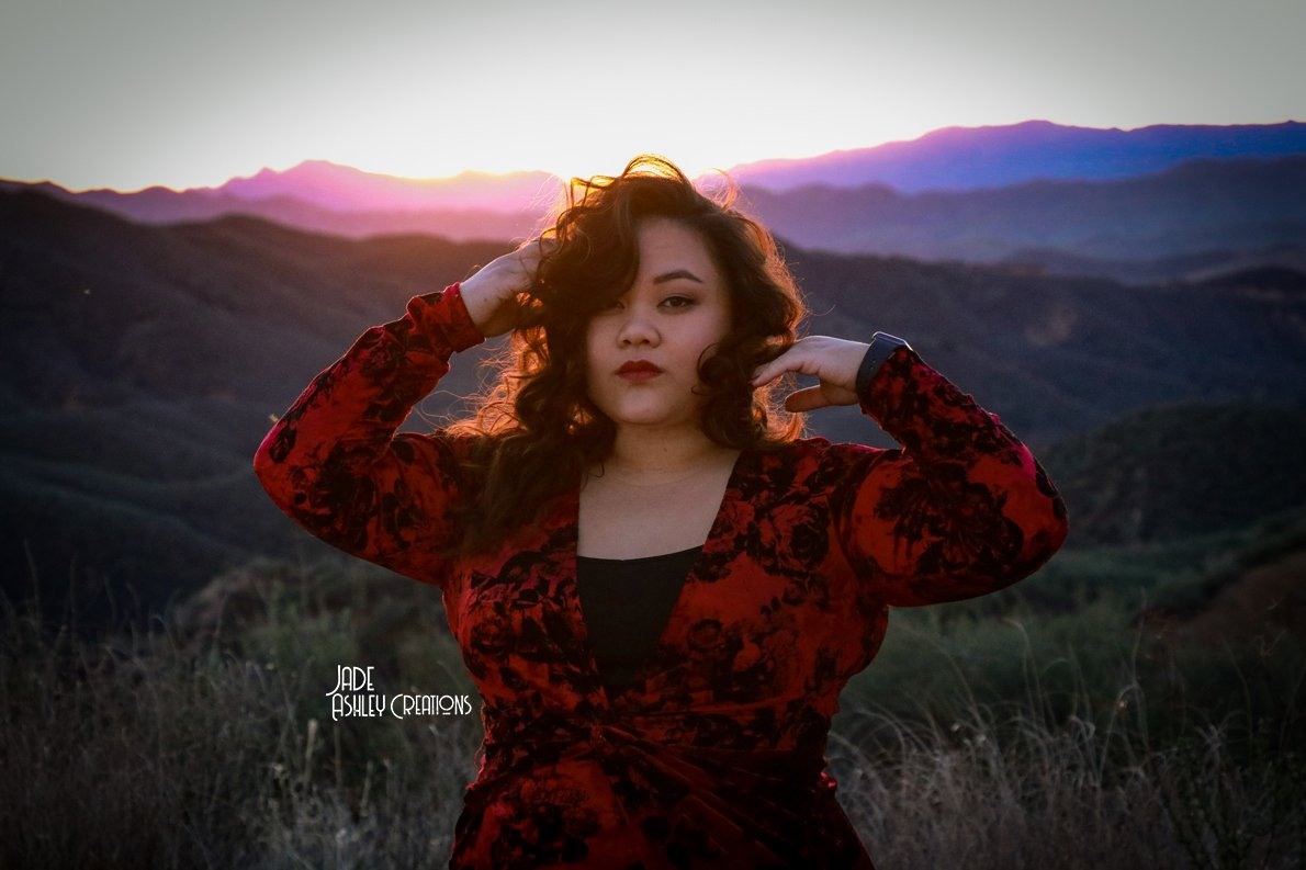A woman with curly hair wearing a red and black dress with mountains and sunset in the background.