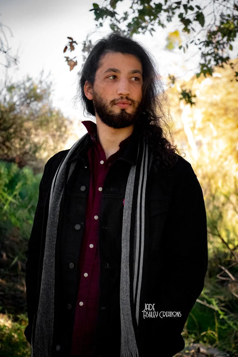 A man with long, dark curly hair and a beard, wearing a black jacket over a maroon shirt with a striped scarf, standing outdoors with trees and sunlight in the background.