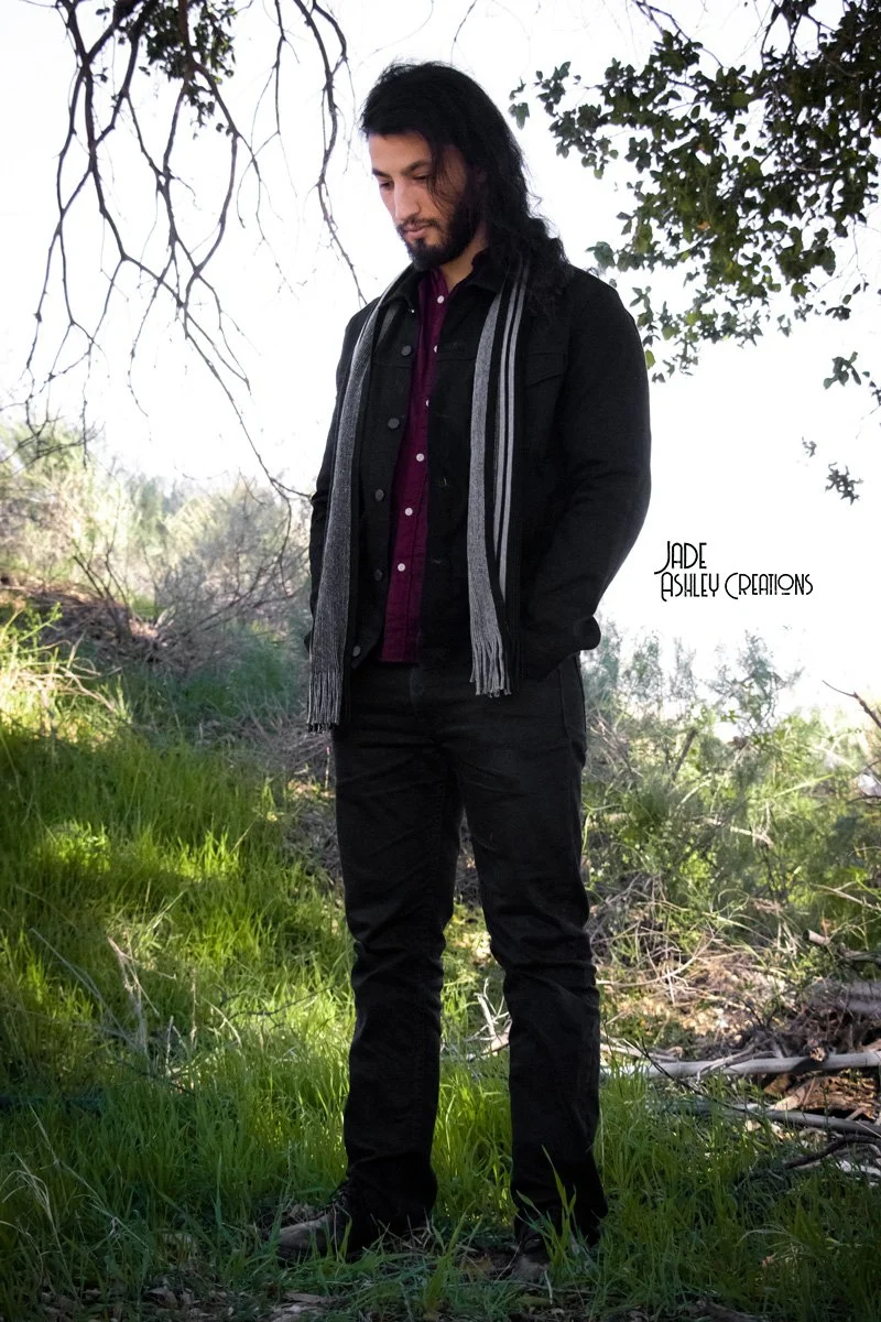 A man with long black hair and a beard standing outdoors in a grassy area with trees and branches overhead. He is wearing a black jacket, a purple shirt, and a gray scarf, looking down with a thoughtful expression.