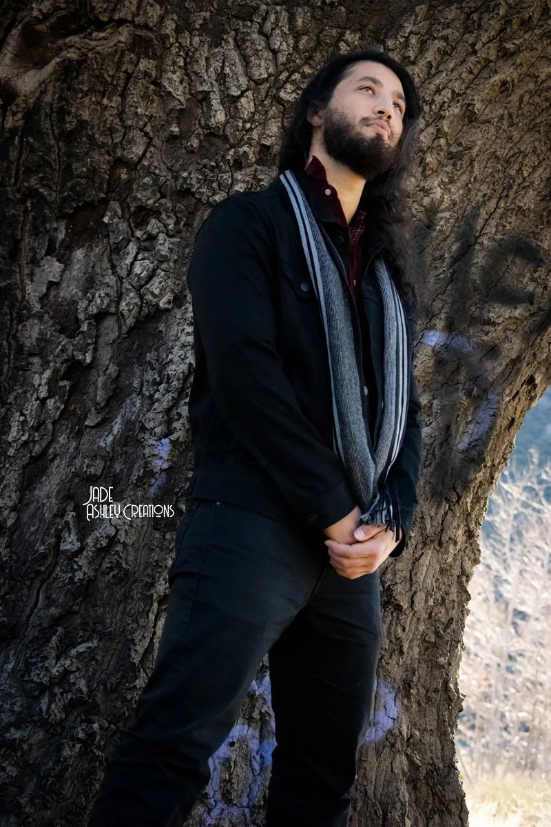 A man with long dark hair and a beard stands outdoors against a large, textured tree trunk, wearing a black jacket and gray scarf, with a contemplative expression.