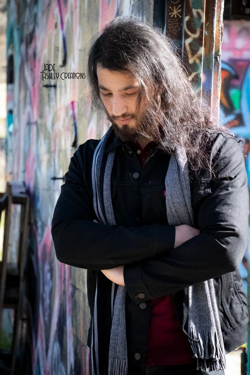 Young man with long hair, closed eyes, wearing a black jacket and patterned scarf, with arms crossed, standing against a colorful graffiti wall.