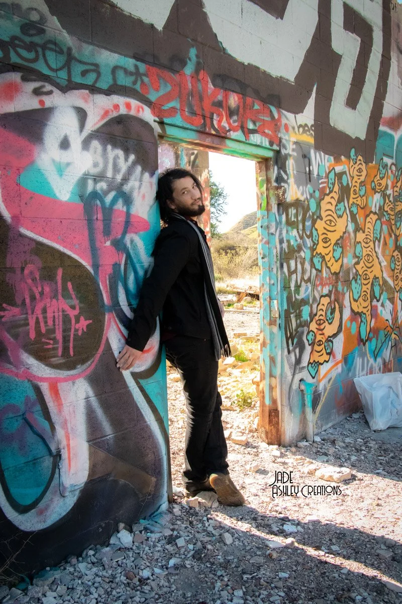 A young man with long hair and a beard standing against a graffiti-covered wall in an abandoned area. The graffiti includes colorful tags and illustrations, with a doorway leading to an outdoor scene with hills and trees.