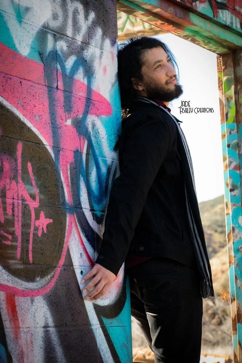 A man with long dark hair and a beard leaning against a graffiti-covered wall in an outdoor setting, with a structure in the background.