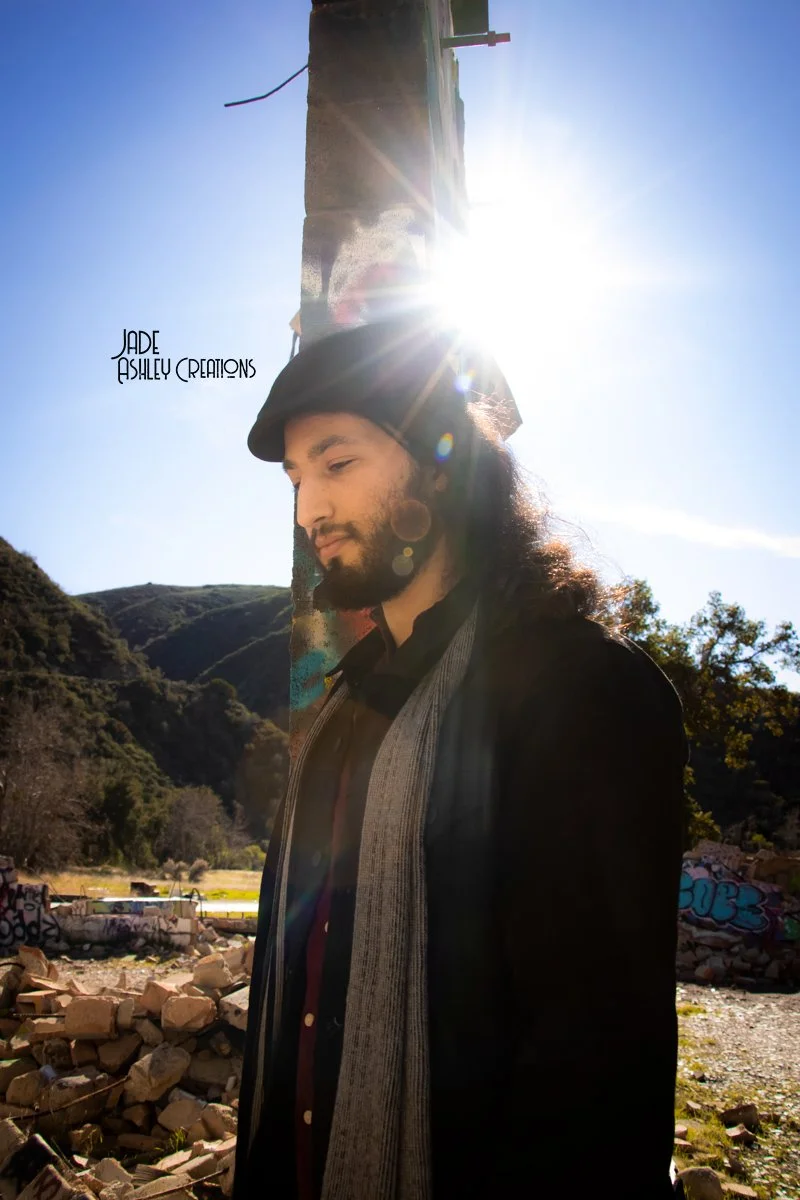 A man with long hair and a beard, wearing a hat and coat, standing outdoors with a bright sun behind him, near a concrete structure and a landscape with hills and graffiti-covered rocks.