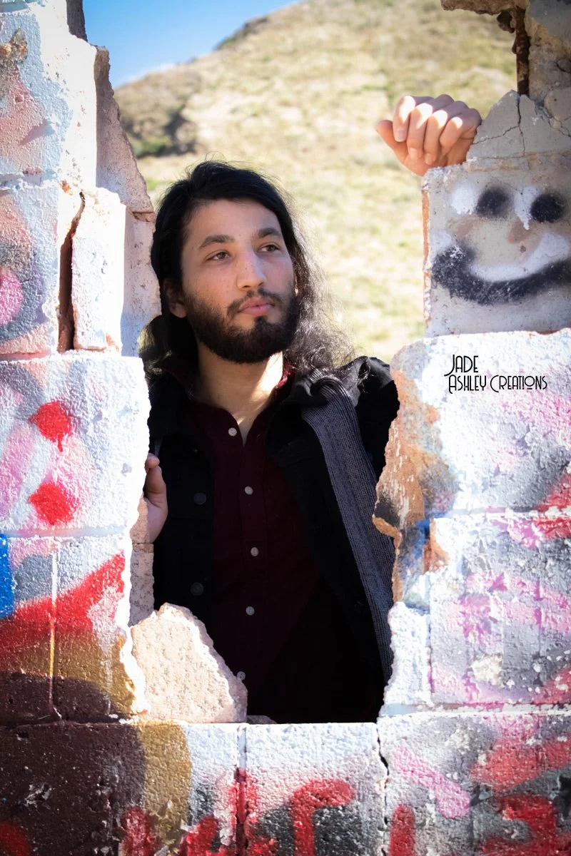 A man with long black hair and a beard looking thoughtfully through a hole in a graffiti-covered brick wall, with a hilly landscape in the background.