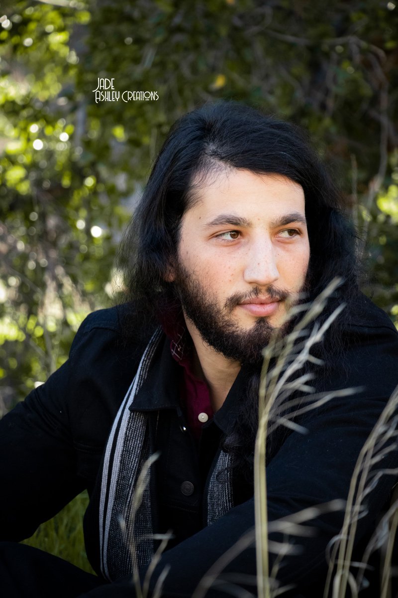 A young man with long dark hair and a beard sitting outdoors in a natural setting with greenery and sunlight.