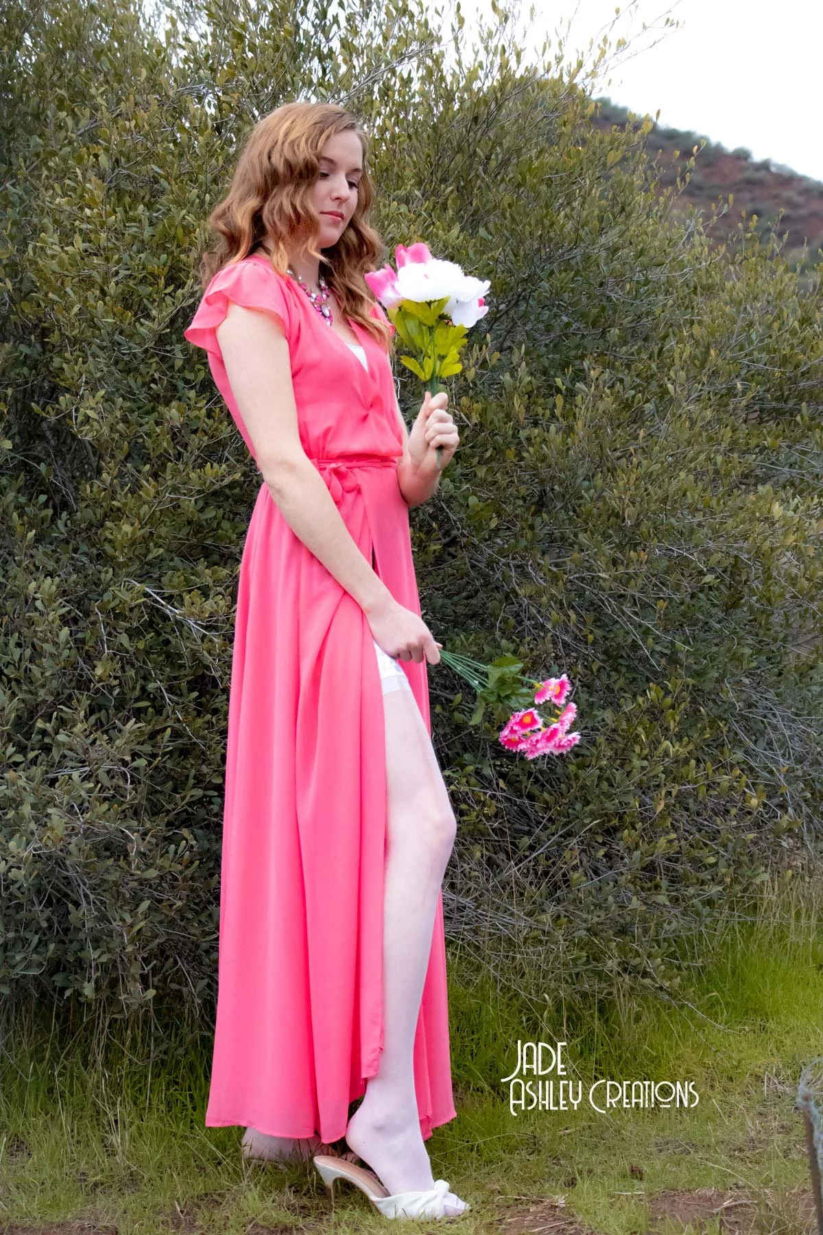 A woman with red hair wearing a bright pink dress standing outdoors in front of dense green bushes, holding a bouquet of pink flowers in one hand and a larger pink and white flower in the other.