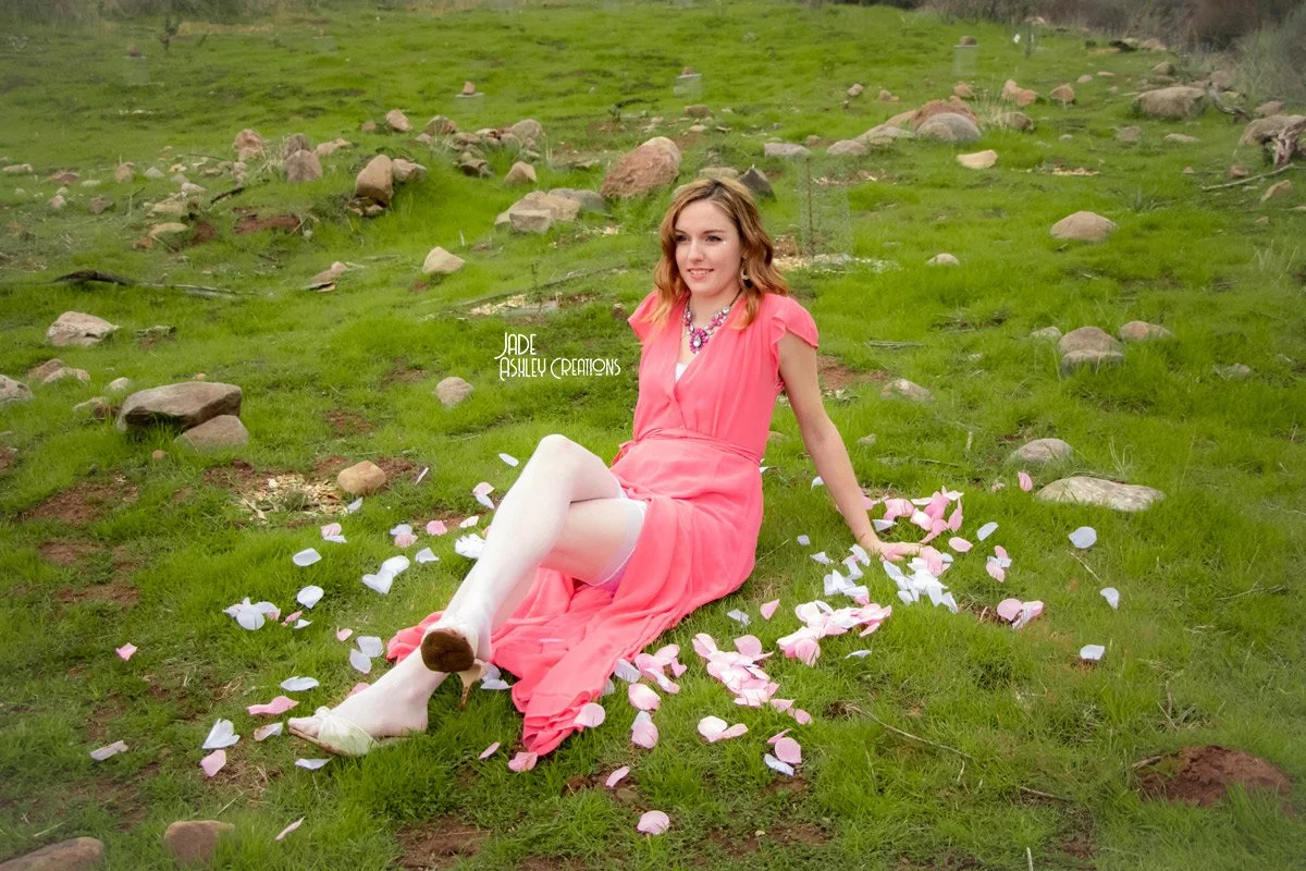 A woman in a pink dress and white tights sitting on a grassy field scattered with pink and white flower petals, with rocks and a few trees in the background.