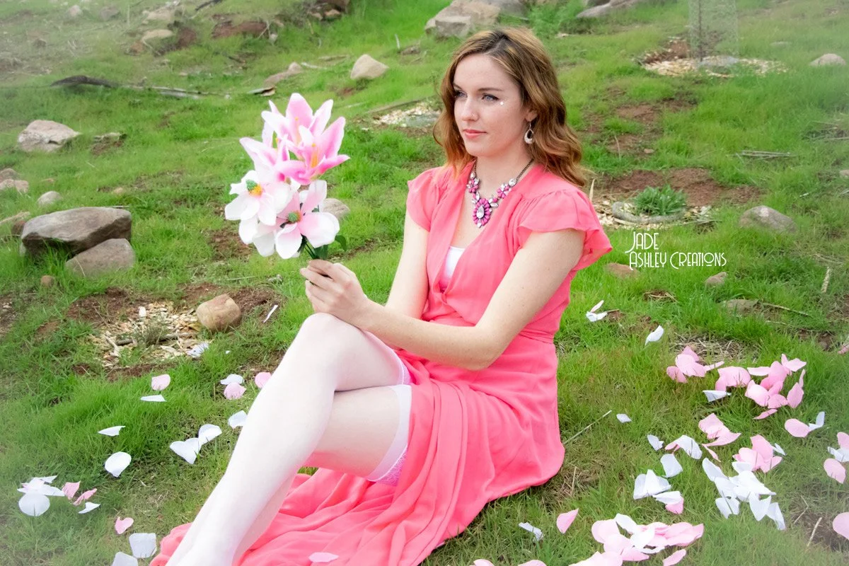 A woman sitting on grass holding a bouquet of large pink and white artificial flowers with scattered petals around her, outdoors in a grassy area with rocks.