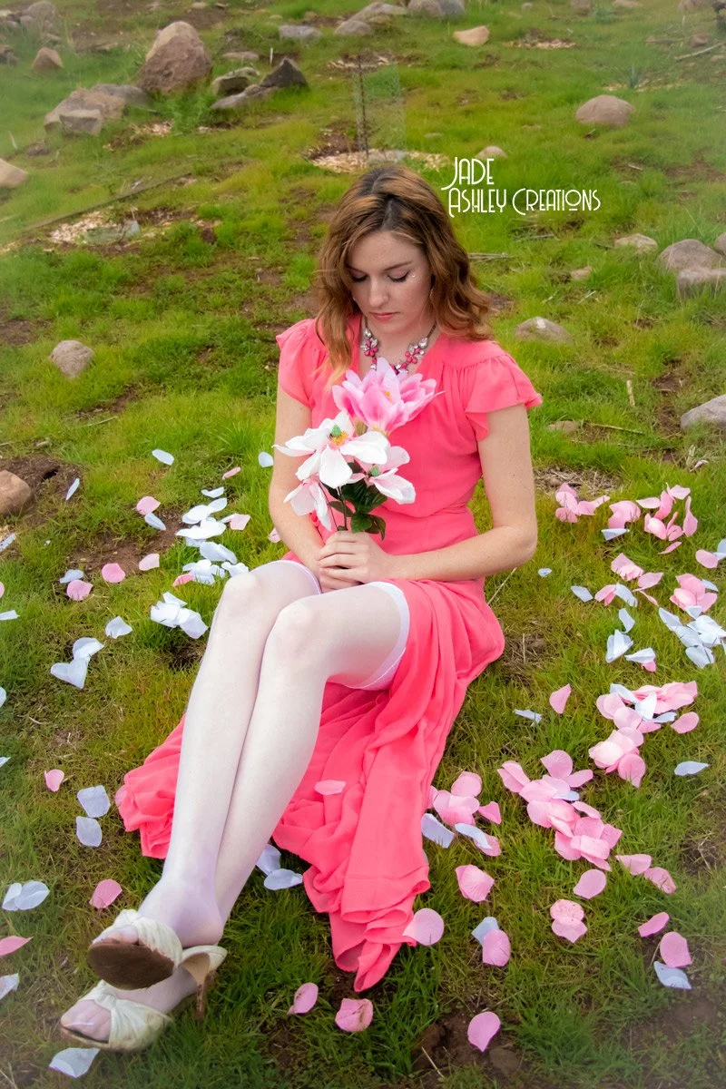 A woman in a pink dress sitting on grass, holding a large flower, surrounded by pink and white petals, with a rocky landscape in the background.