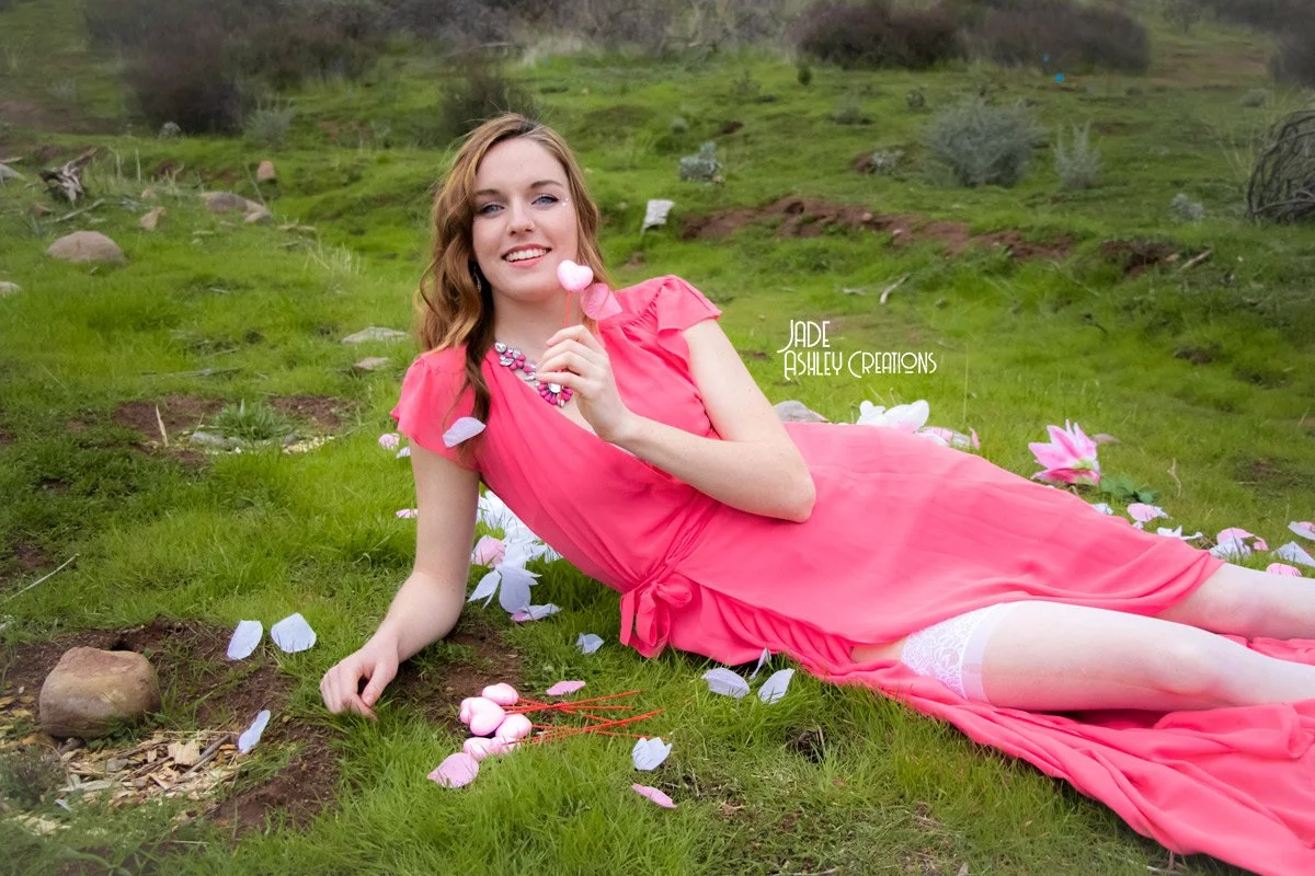 A young woman in a pink dress lying on grass in a field with pink and white flower petals scattered around, holding a pink lollipop and smiling.