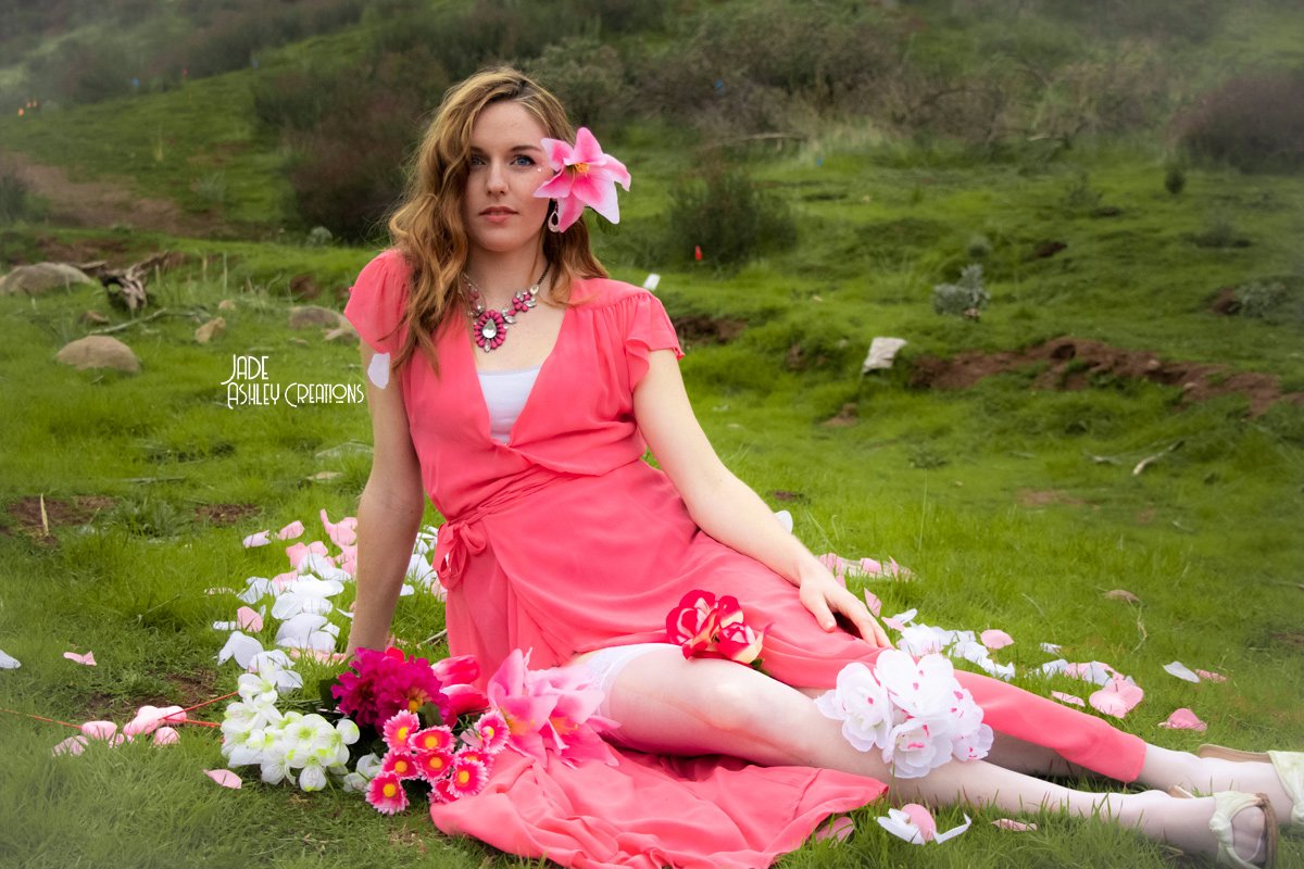 A woman in a pink dress sitting on a grassy field surrounded by pink and white flowers, with a hill and trees in the background.
