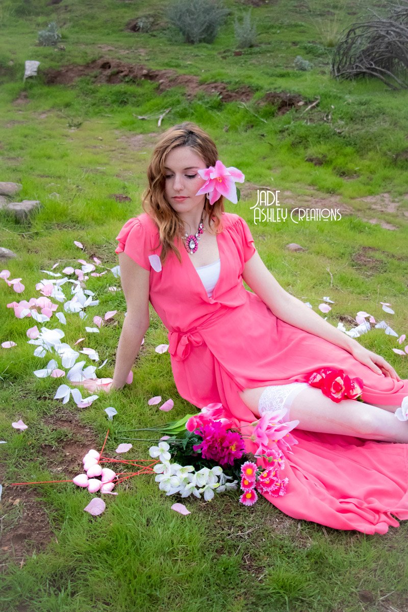 A young woman with wavy brown hair sits on green grass, wearing a pink dress and white thigh-high stockings. She has a large pink flower in her hair, a pink flower necklace, and is surrounded by scattered pink and white flower petals and a bouquet of