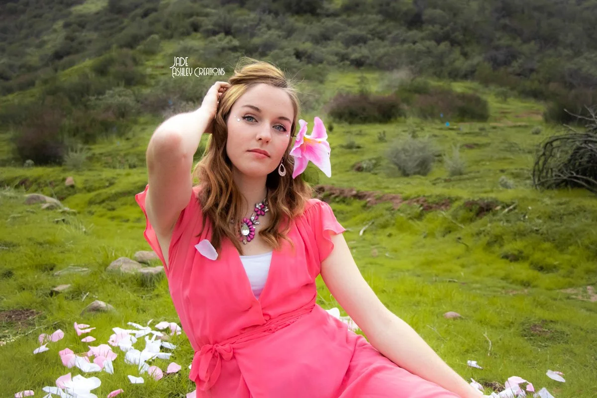 A woman in a pink dress sitting on grass with a pink lily in her hair, surrounded by flower petals in a green, hilly landscape.