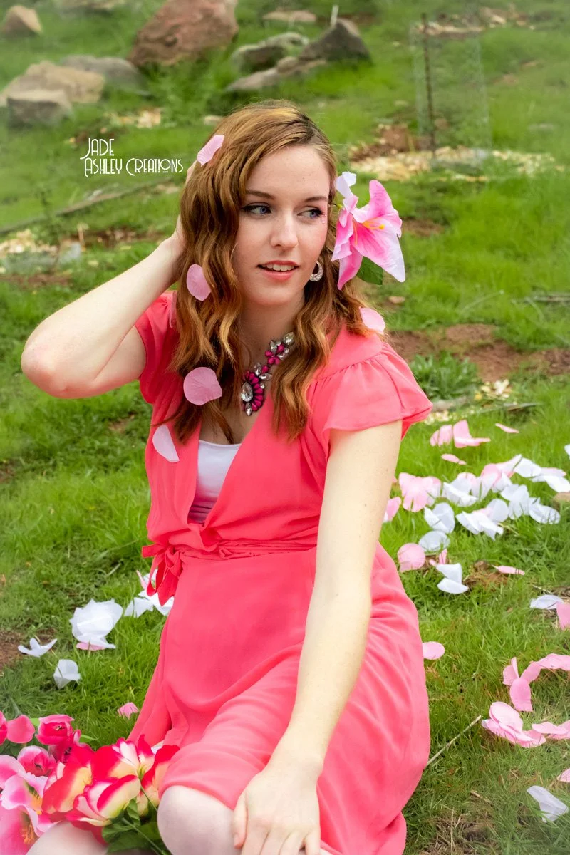 A woman with wavy, auburn hair outdoors, sitting on green grass amidst pink and white flower petals, wearing a coral pink dress and a statement necklace.