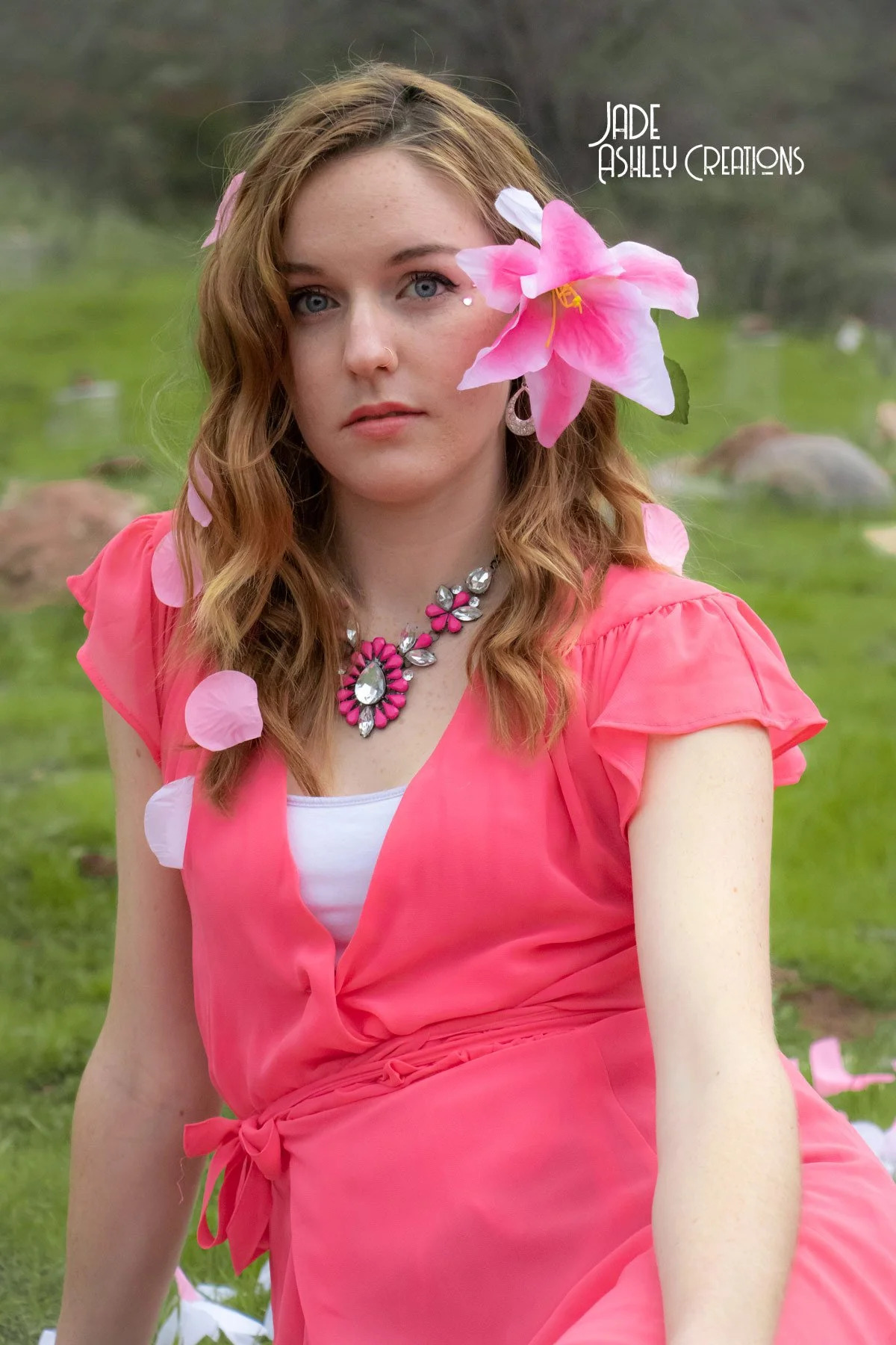 A young woman with red hair wearing a pink dress and necklace, with a large pink flower near her face, sitting outdoors on grass with blurred nature background.