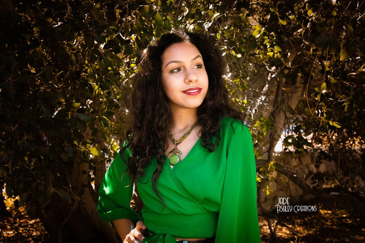 A young woman with long curly dark hair wearing a bright green blouse and a statement necklace, posing outdoors in front of a bush or tree with sunlight filtering through the leaves.
