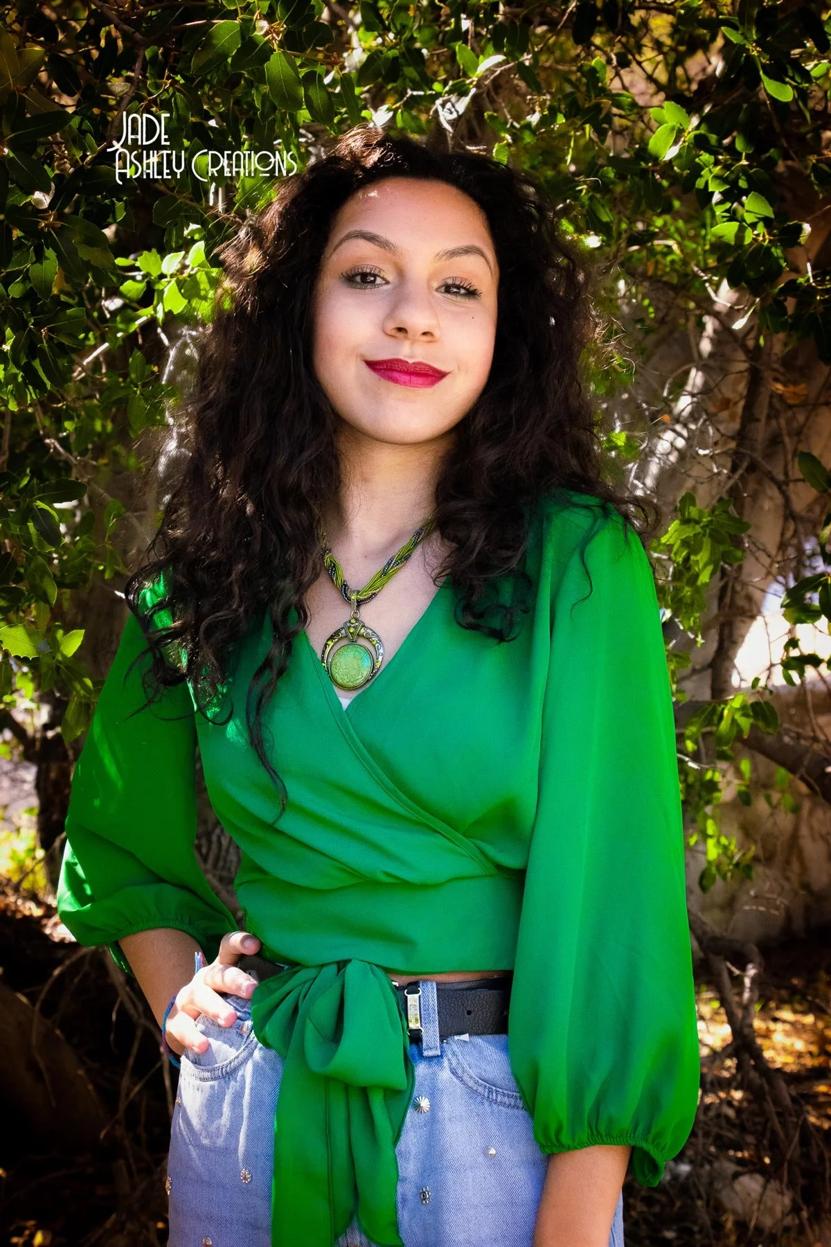 A woman with curly dark hair and red lipstick is smiling outdoors, wearing a bright green wrap top, light blue jeans with embellishments, and a green scarf tied at her waist. She has a layered necklace with a green pendant, standing in front of leafy