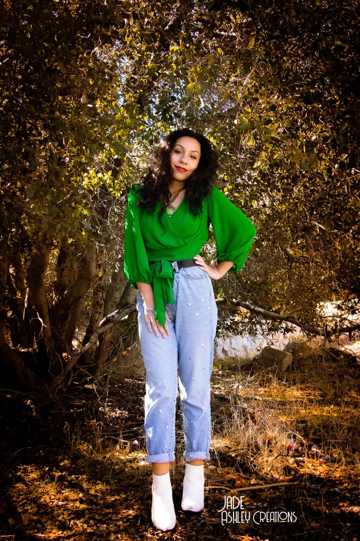 A woman in a green wrap blouse, light blue jeans with embellishments, and white boots, standing outdoors in front of dense trees with sunlight filtering through the leaves.