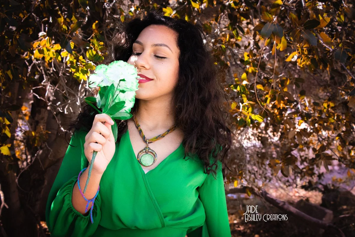 A woman with dark, curly hair is wearing a green blouse and a colorful necklace. She is holding and smelling a green and white flower. The background is filled with brown and yellow leaves.