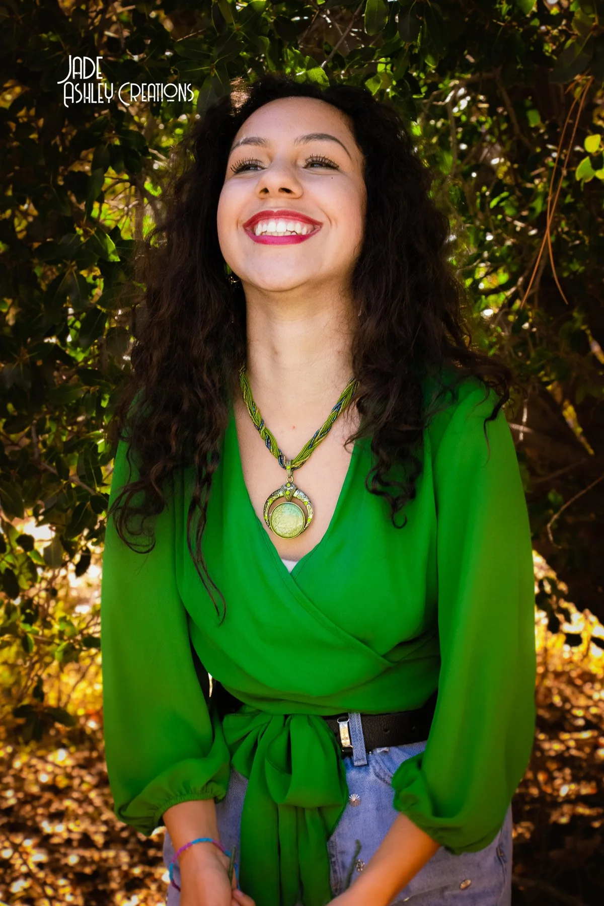 A woman in a vibrant green blouse stands outdoors in front of leafy greenery, smiling happily.