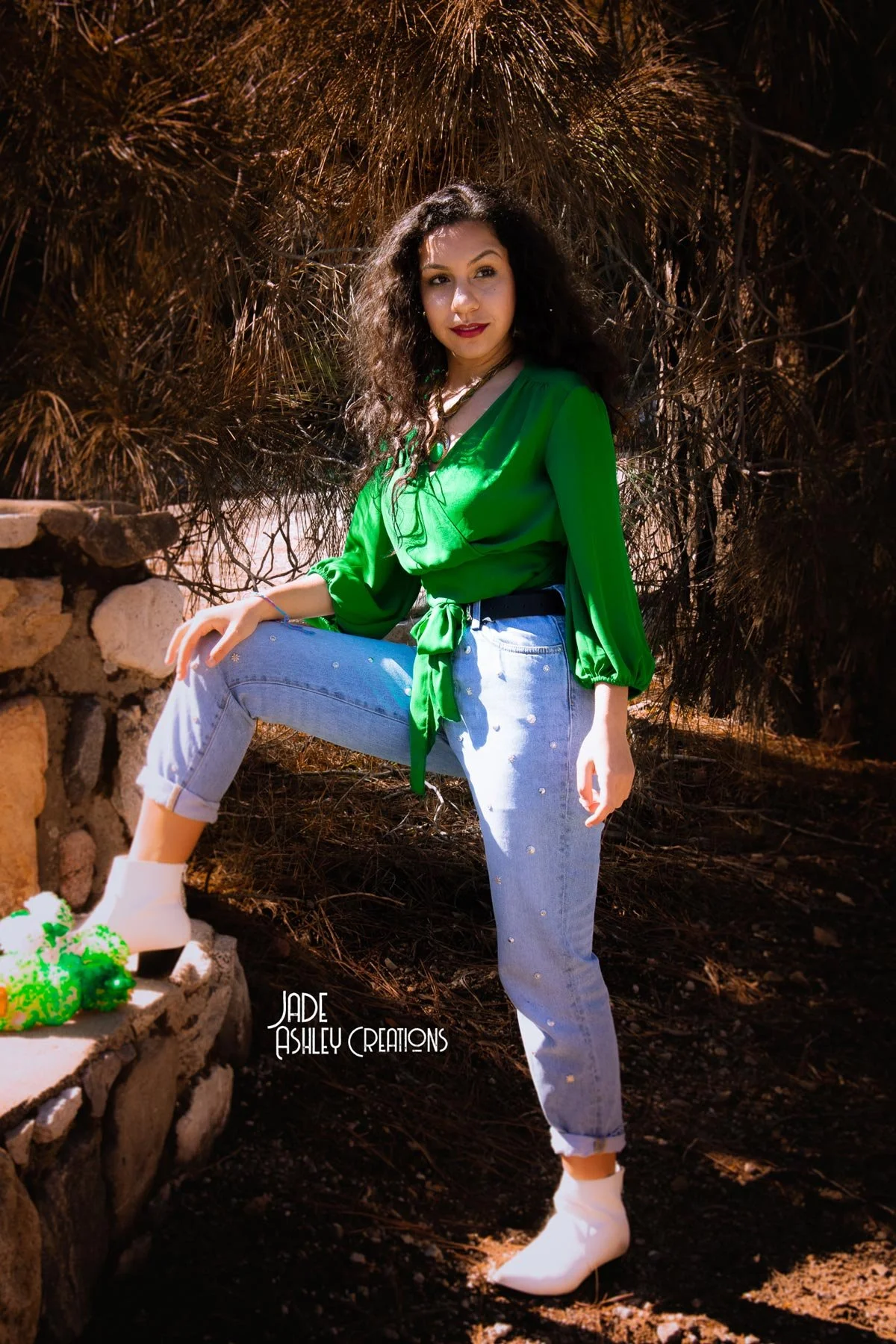 A woman with curly dark hair wearing a green blouse, light blue jeans with embellishments, white boots, sitting outdoors near a stone wall and dense bushes.