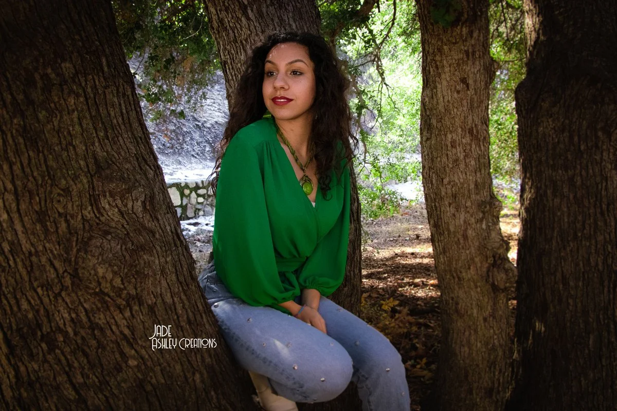 A woman with dark curly hair sitting between three trees in a wooded area, wearing a green blouse and jeans, with green jewelry, looking to the side.