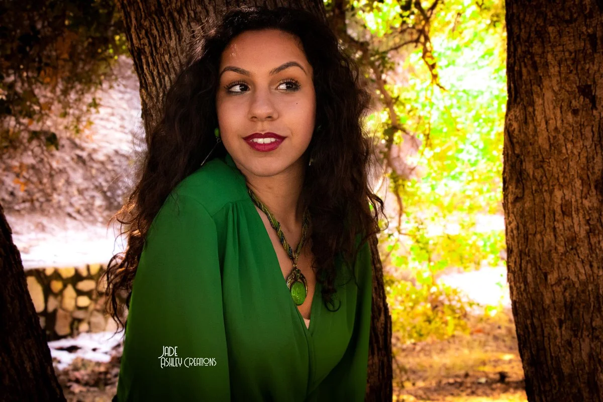 A woman with dark curly hair and red lipstick wearing a green blouse and necklace, leaning against a tree in a park or forest with colorful autumn leaves in the background.