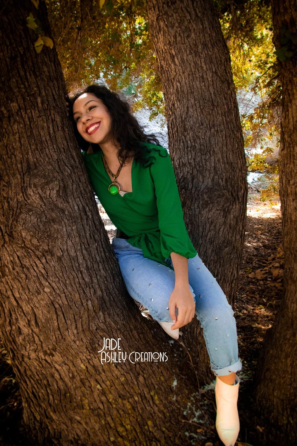 A woman in a green blouse and jeans with embellishments, sitting between tree trunks outdoors, smiling and leaning her head against a tree.
