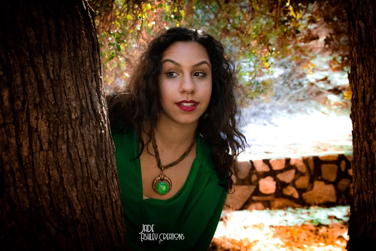 A woman with long, curly dark hair wearing a green top and a large necklace with a green pendant. She is outdoors between two trees, with a background of autumn leaves and a stone wall.