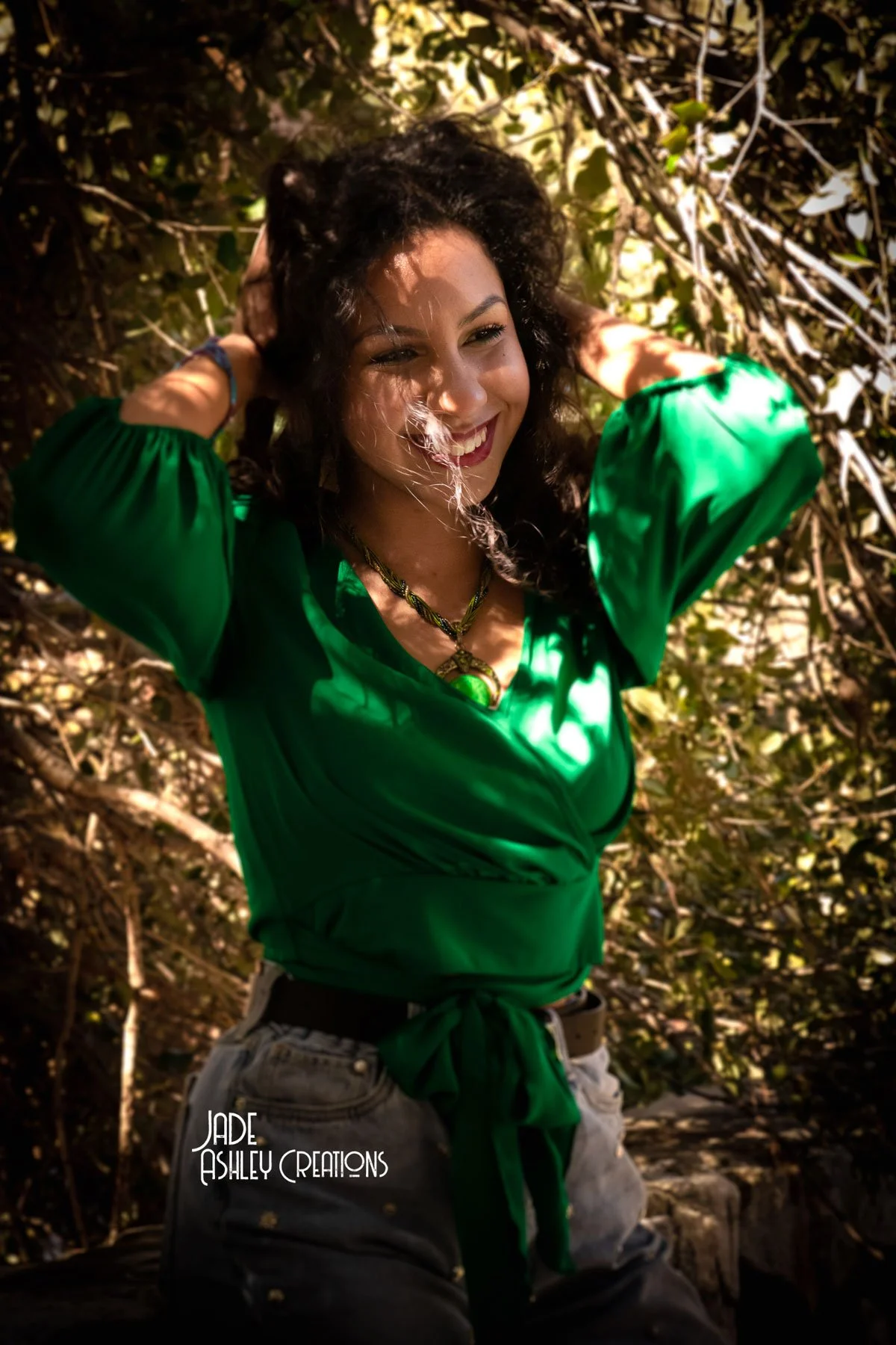 A woman with curly dark hair smiling with her eyes closed, wearing a green blouse and jewelry, posing outdoors in front of bushes with sunlight filtering through the leaves.
