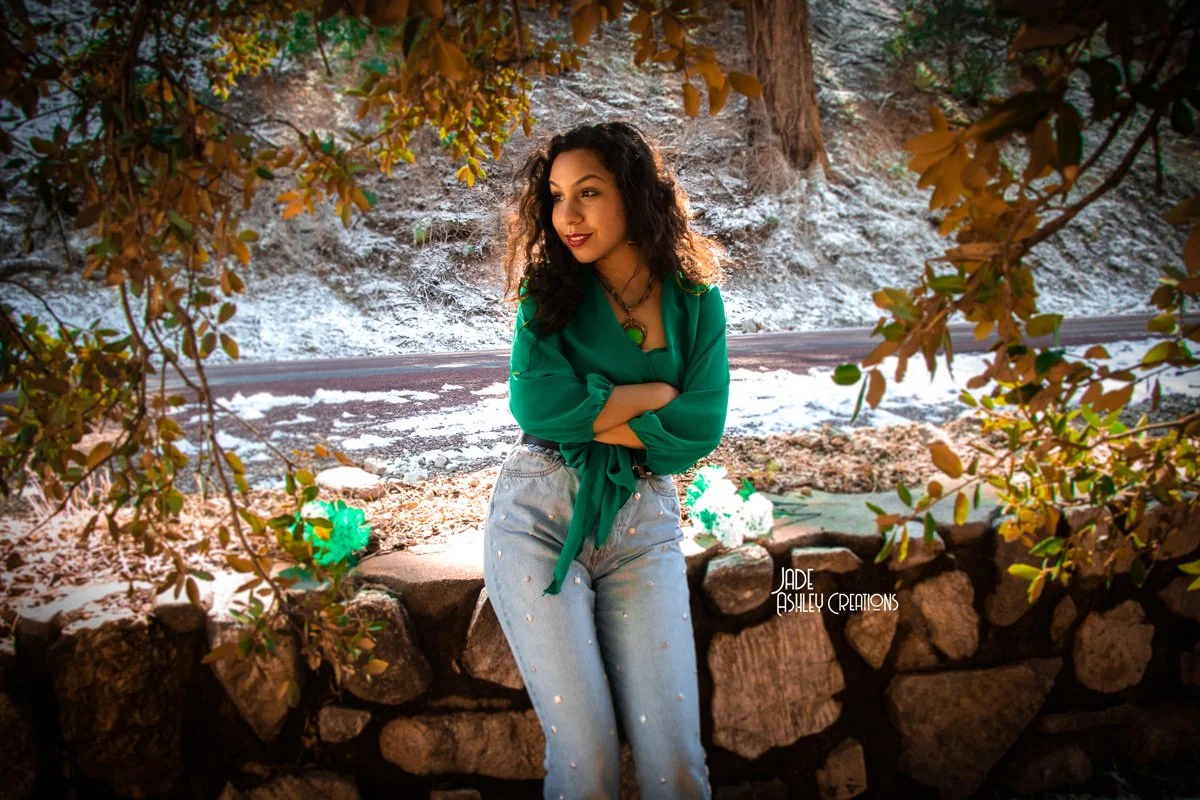 Woman with curly hair in a green blouse and light jeans standing outdoors behind a stone wall, surrounded by leaves, with snow on the ground and a road in the background.