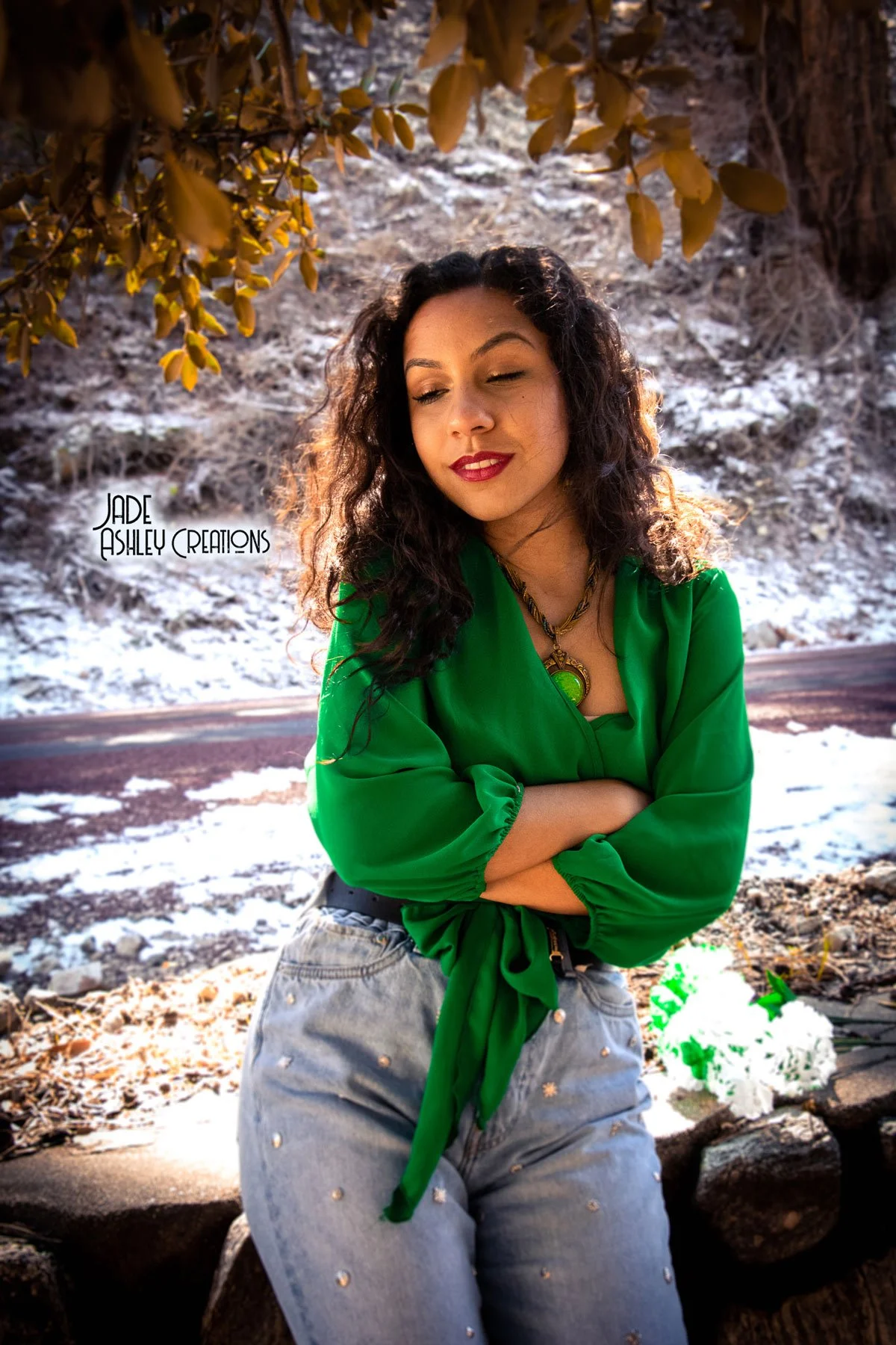 A woman with curly dark hair and red lipstick stands outdoors with a snowy backdrop, wearing a bright green blouse and jeans with embellishments. She has her arms crossed and a pendant necklace, and she looks contemplatively to the side.