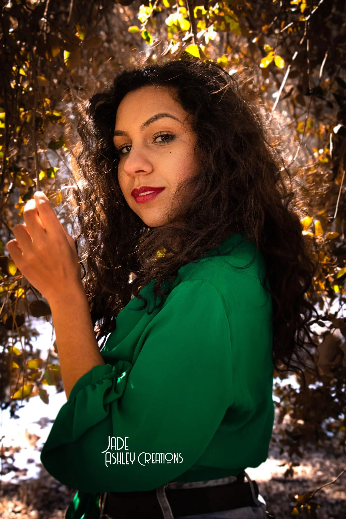 A woman with curly dark hair and red lipstick standing outdoors among brown and yellow leaves, wearing a green top, looking over her shoulder at the camera.