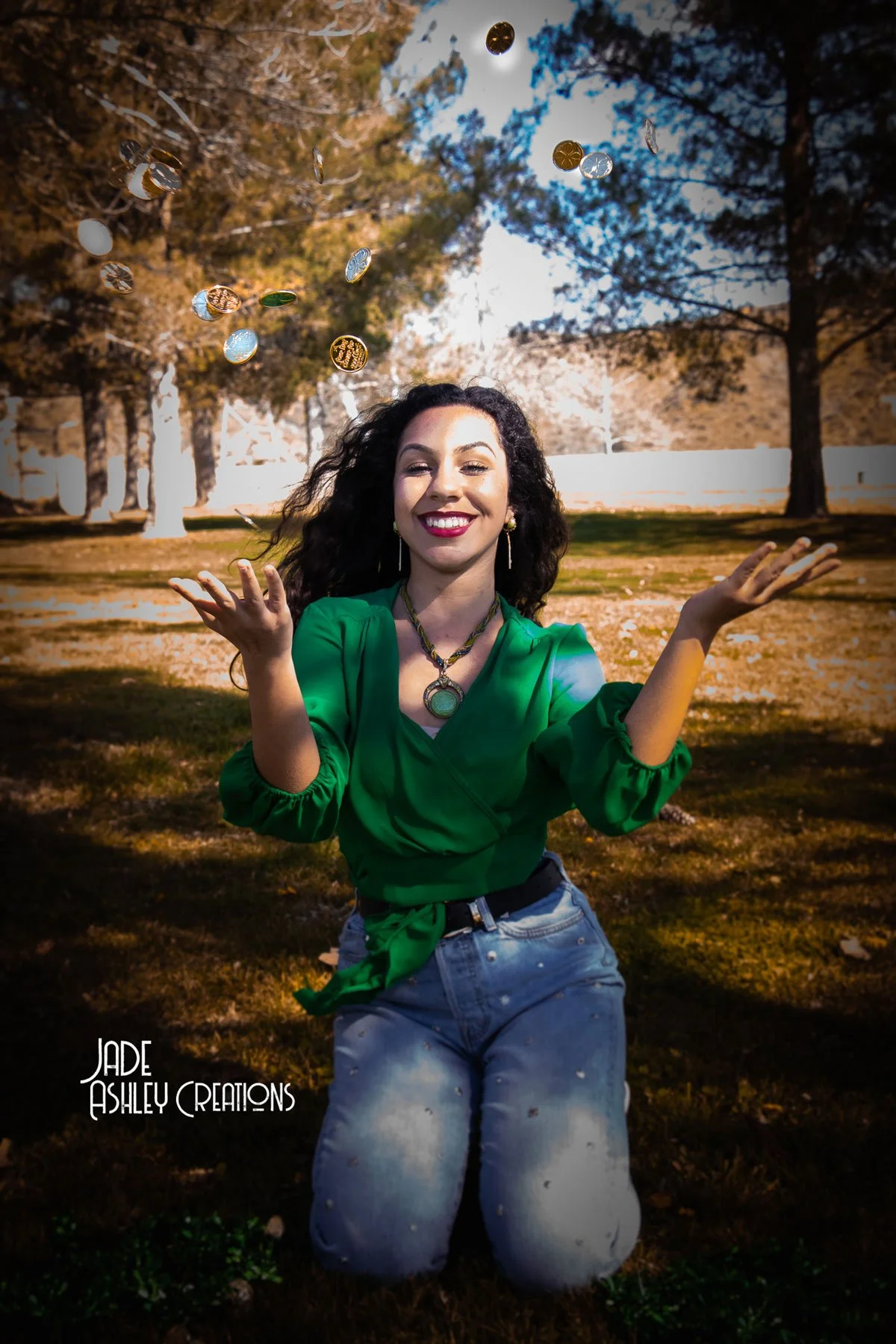 A woman with curly dark hair smiling, kneeling on grass with trees in the background, throwing confetti in the air. She is wearing a green blouse, blue jeans, and jewelry, and the photo is taken outdoors during daytime.