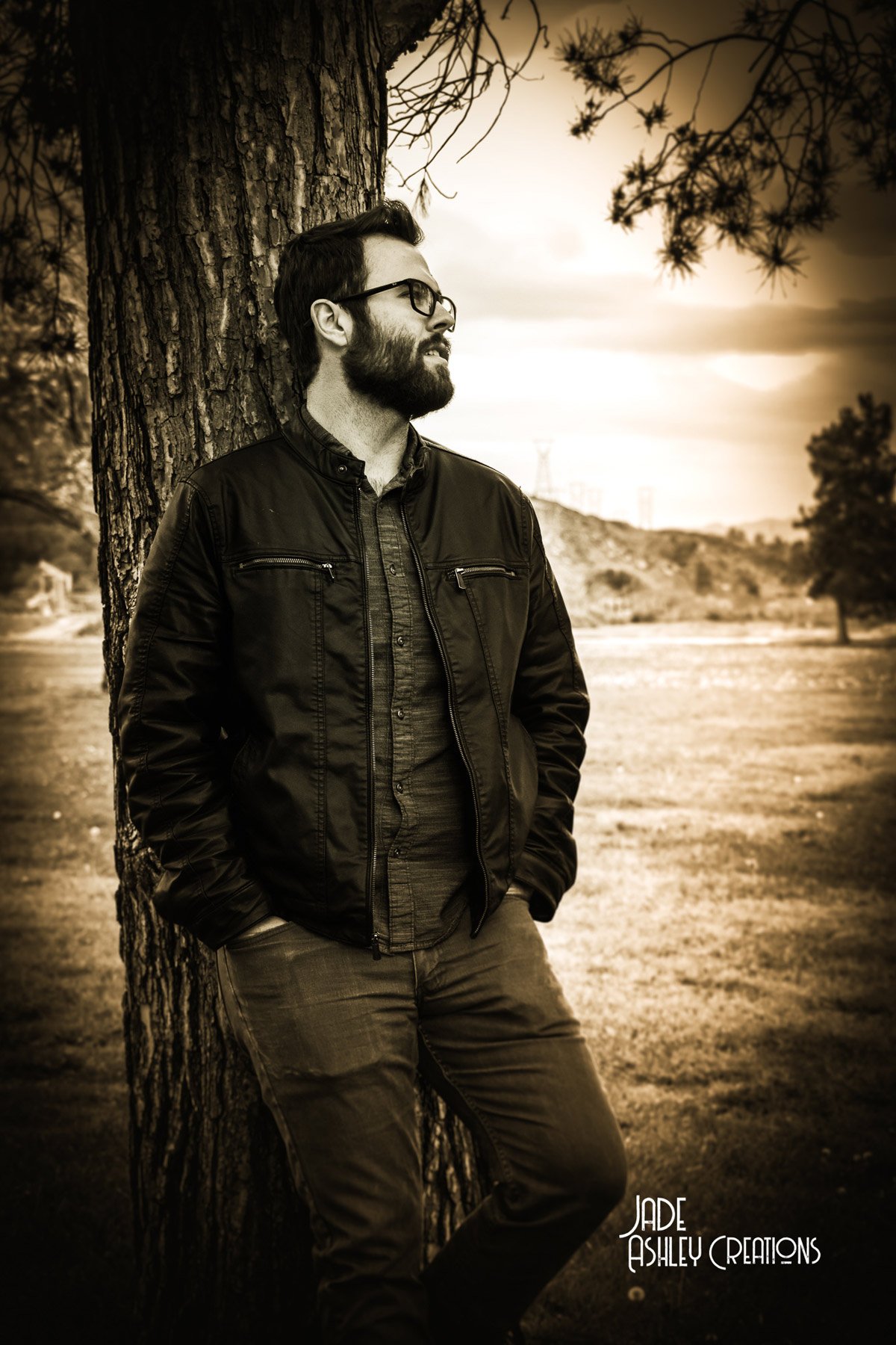 A man with a beard and glasses leaning against a tree outdoors during sunset, with a rural landscape and hills in the background.
