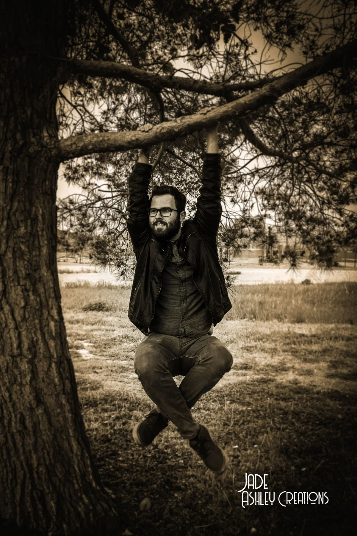 A man hanging from a tree branch, with his hands gripping the branch and legs crossed, smiling and looking to the side, in a sepia tone photograph.